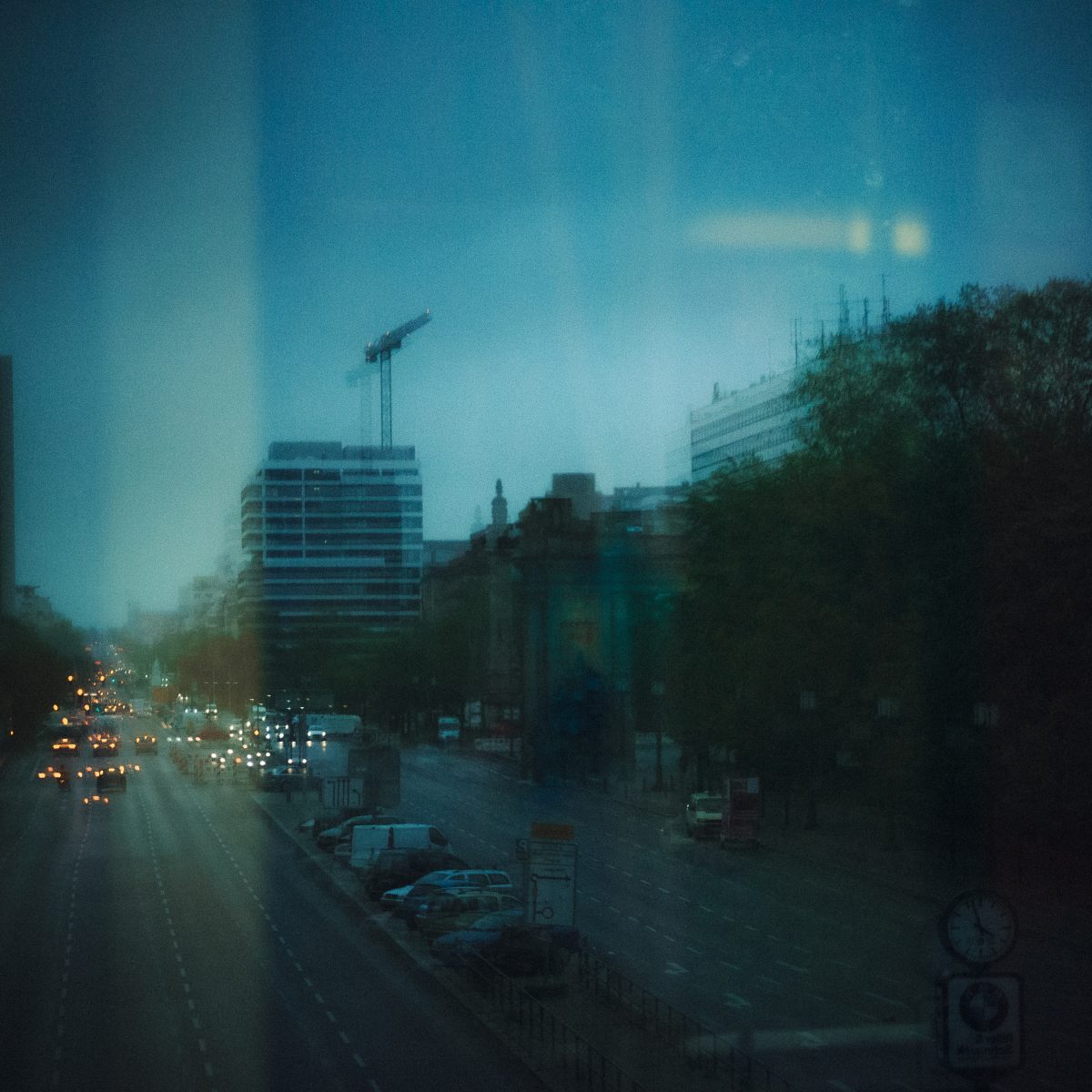 Layered reflection of a Berlin boulevard at blue hour, seen through glass with ghosting and a construction crane