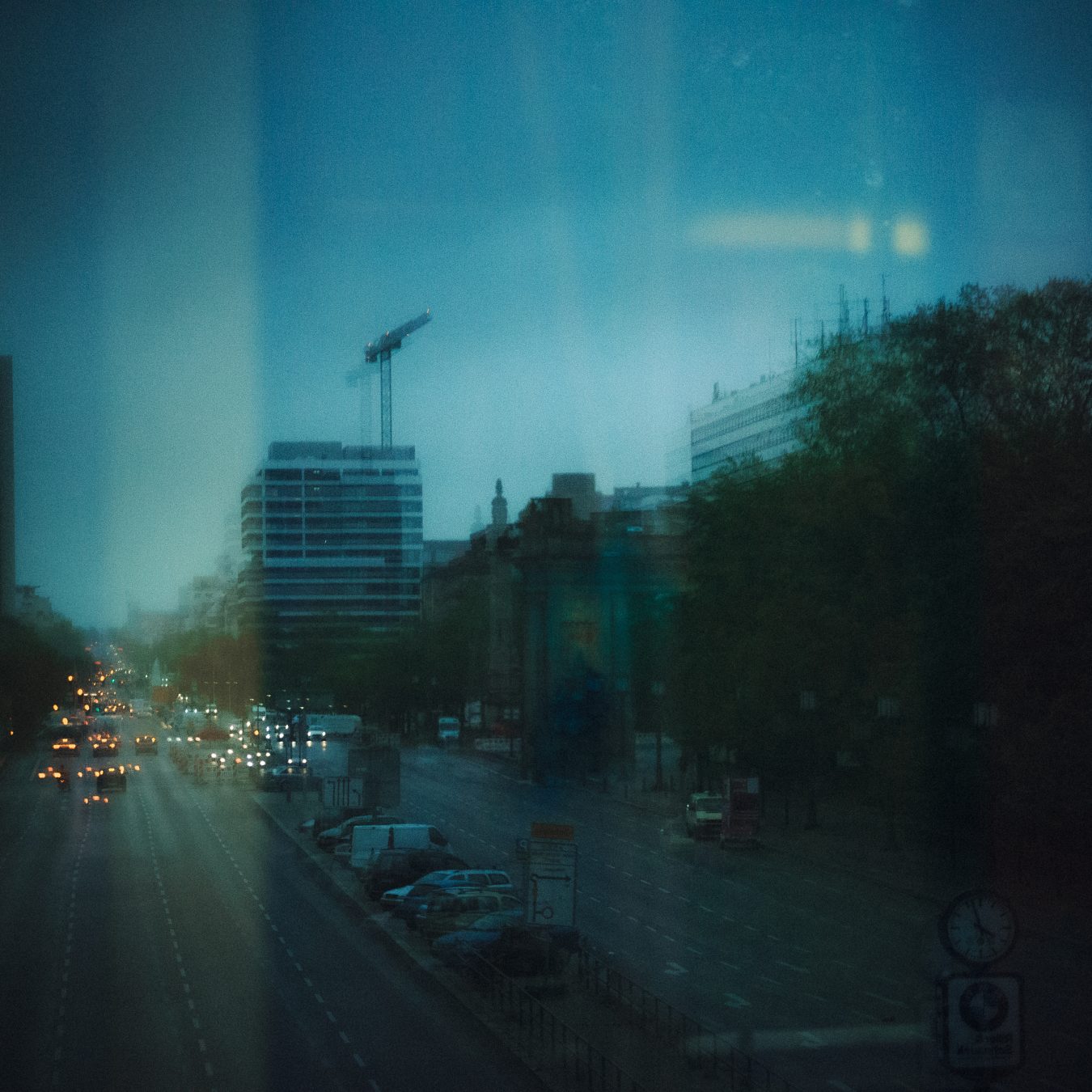 Layered reflection of a Berlin boulevard at blue hour, seen through glass with ghosting and a construction crane