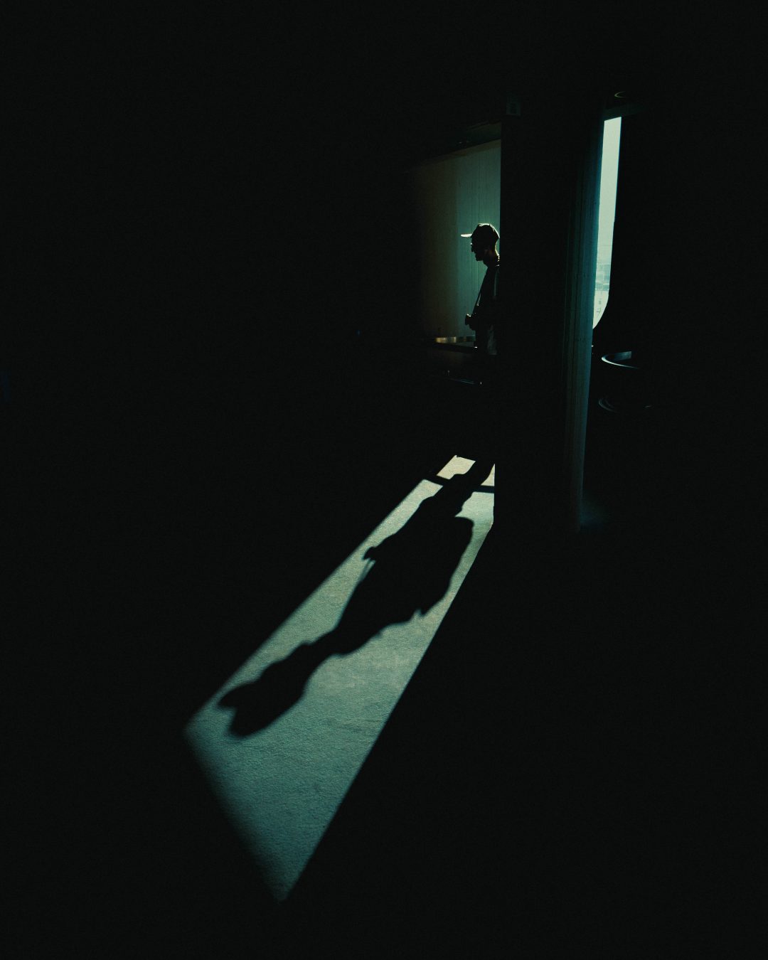 Silhouetted figure with a camera inside a dark interior beside a light shaft, casting a long shadow across the floor