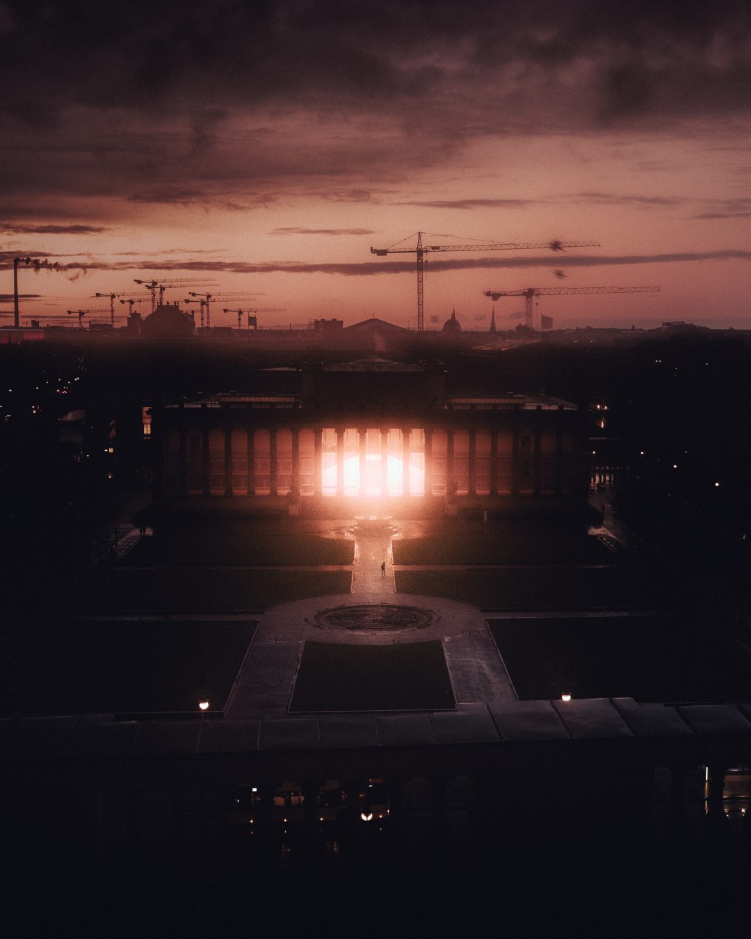 A colonnaded building glowing with light against a dramatic dusk sky, a solitary figure walking a symmetrical path toward it