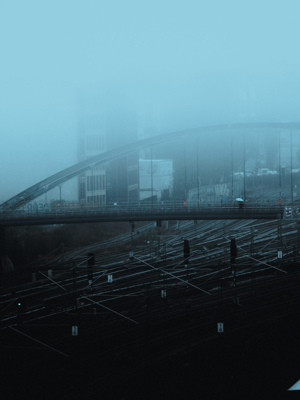 Foggy blue railway bridge over a maze of train tracks, a tiny figure with a white umbrella crossing