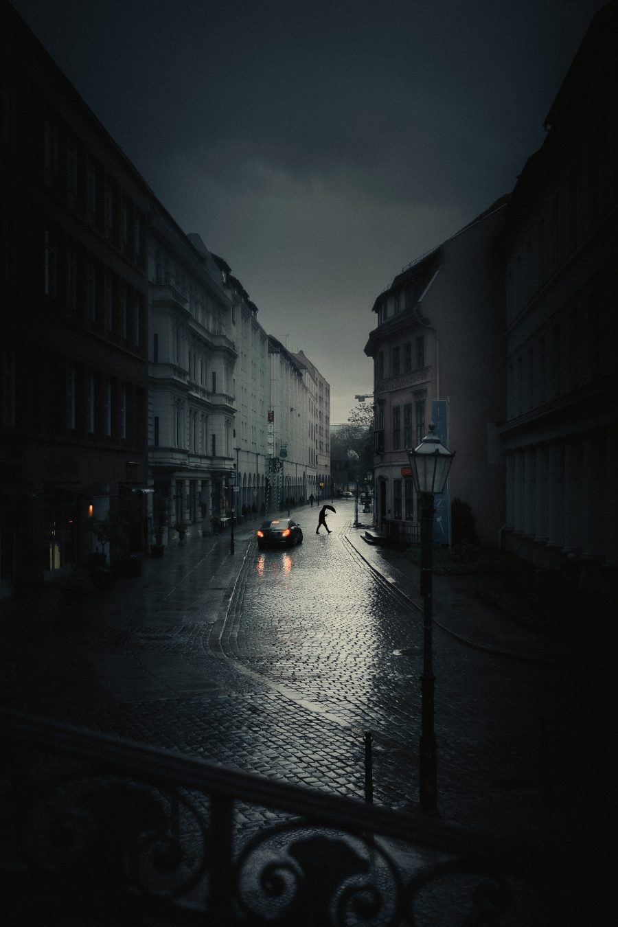 A silhouetted figure with an umbrella crossing a narrow dark cobblestone alley, car taillights and streetlamp glow