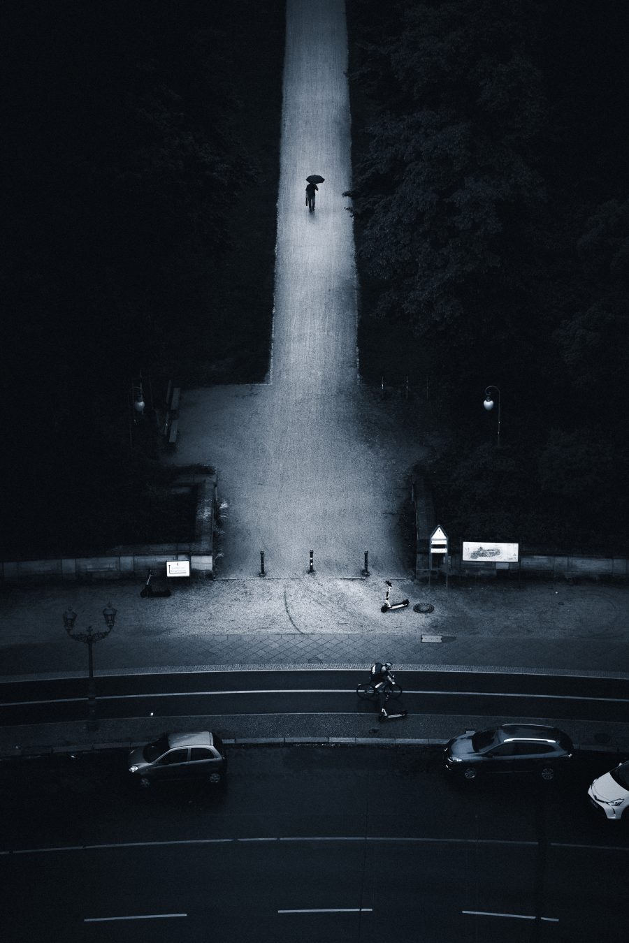 A symmetrical illuminated path cutting through dark trees, a figure with an umbrella walking toward the camera