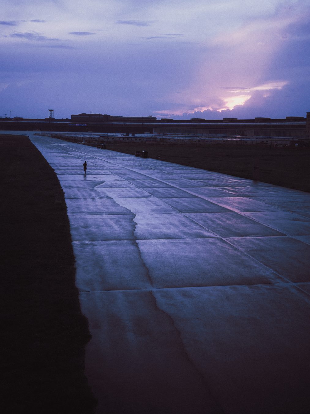 A lone figure walking a wide runway under a violet storm sky split by a beam of light